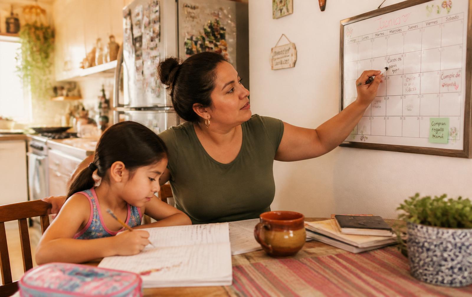 Mamá mexicana organizando el calendario familiar en la cocina mientras su hija hace la tarea